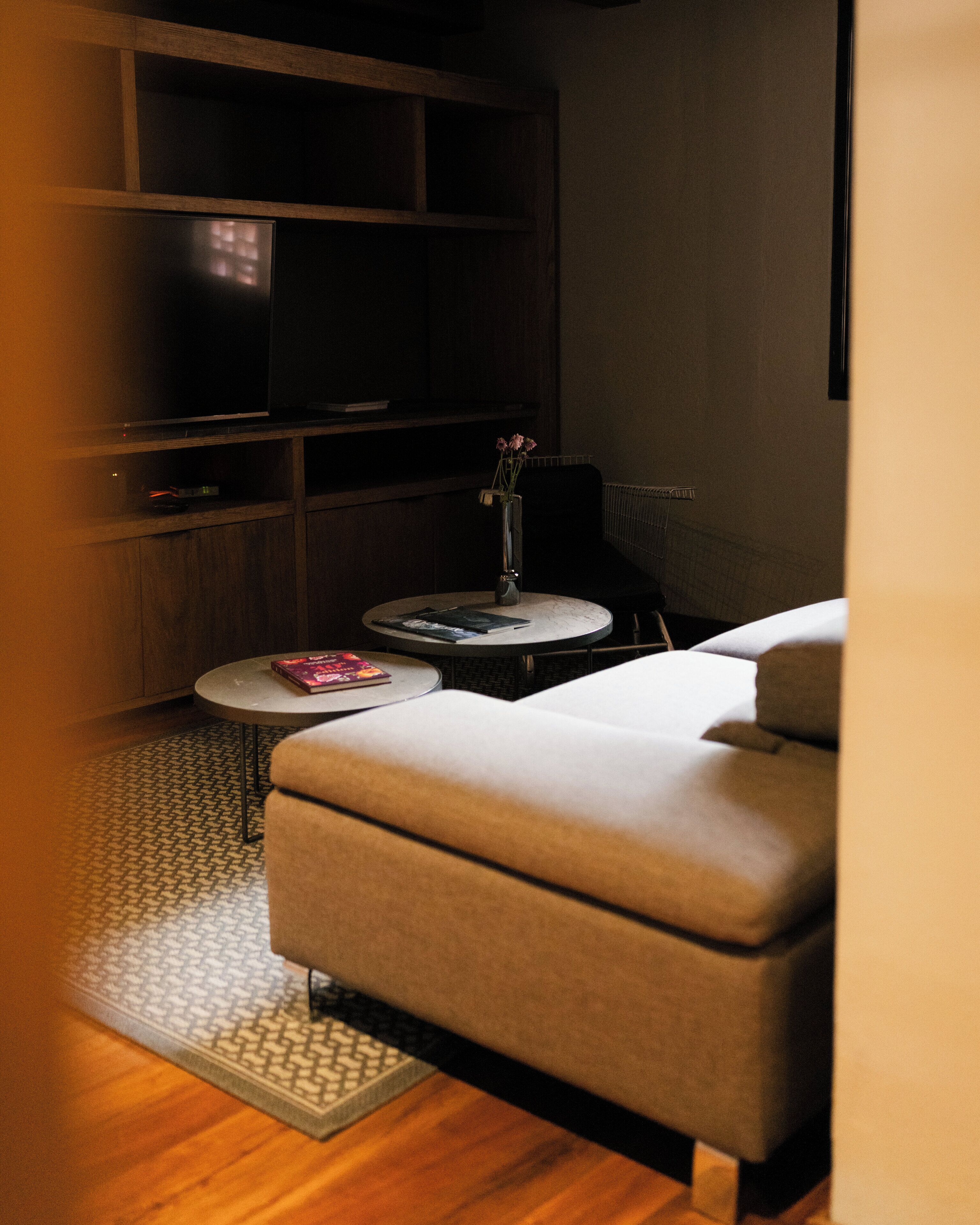 A modern living room with a sofa, a small round table with a book, a vase with flowers, and a television on a shelf.
