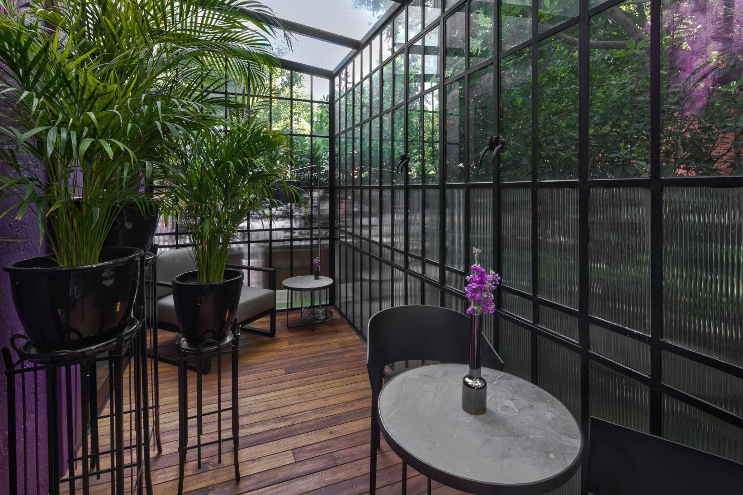 A conservatory with a wooden floor, black metal chairs, a round table, and potted plants.