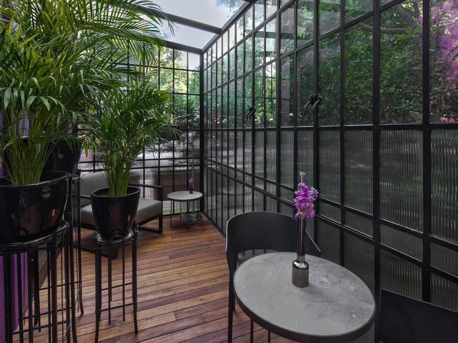 A conservatory with a wooden floor, black metal chairs, a round table, and potted plants.