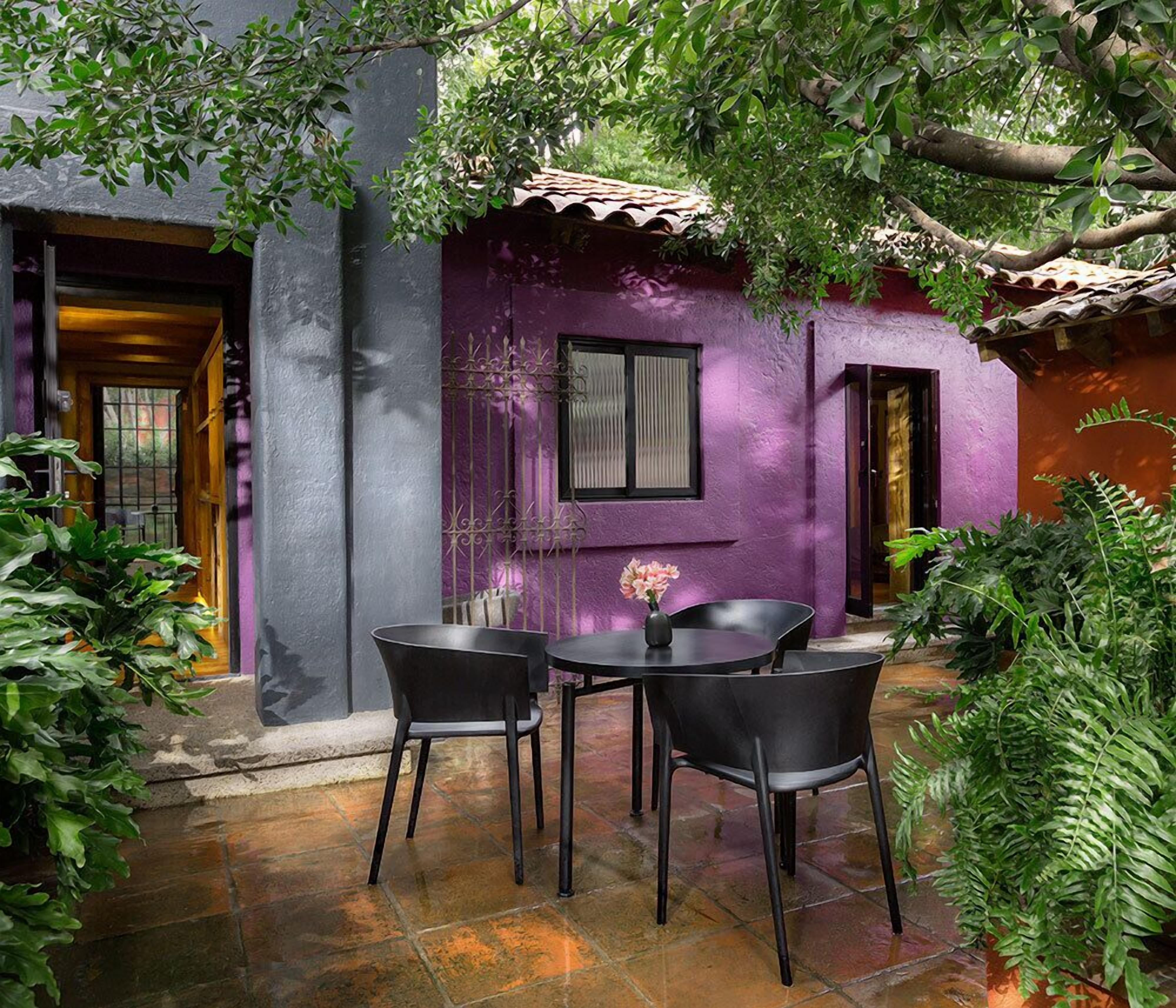 Um pátio externo com duas cadeiras pretas e uma mesa, cercado por vegetação exuberante e um edifício roxo com telhado de azulejos.