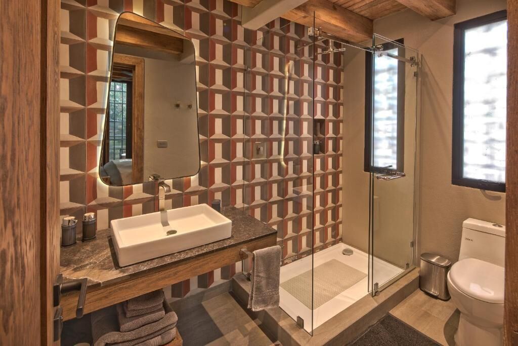 Un bany modern amb un mur amb dibuixos geomètrics, un mirall gran, un lavabo blanc i una dutxa amb envà.