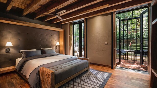 A bedroom with a large bed, wooden headboard, two bedside lamps, and a view of an outdoor patio with a table and chairs. - Morada La Noria (Puebla)