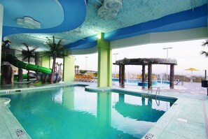 Swimmers can enjoy the pool surrounded by a deck and palm trees, with a green slide for kids and a waterfall feature nearby.