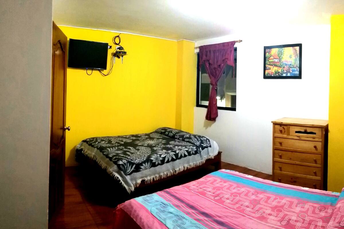 Quarto com cama, cómoda de madeira, televisão na parede e janela com cortinas.