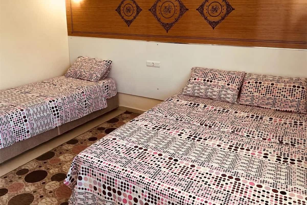 A room with two beds, each with patterned bedding, a wooden headboard with decorative elements, and a light switch on the wall.