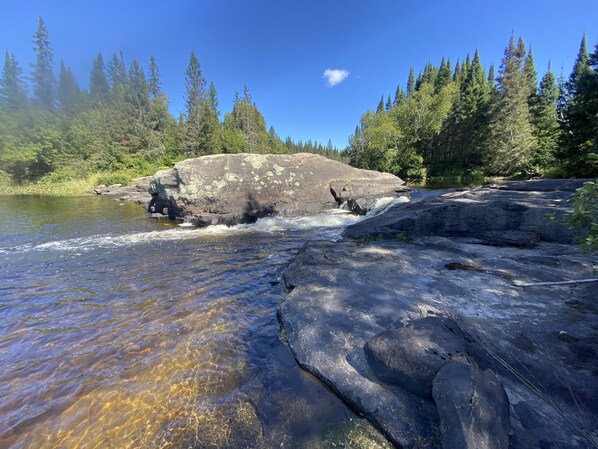 Un río fluye sobre rocas con agua cristalina, rodeado de un bosque denso y una gran formación rocosa.