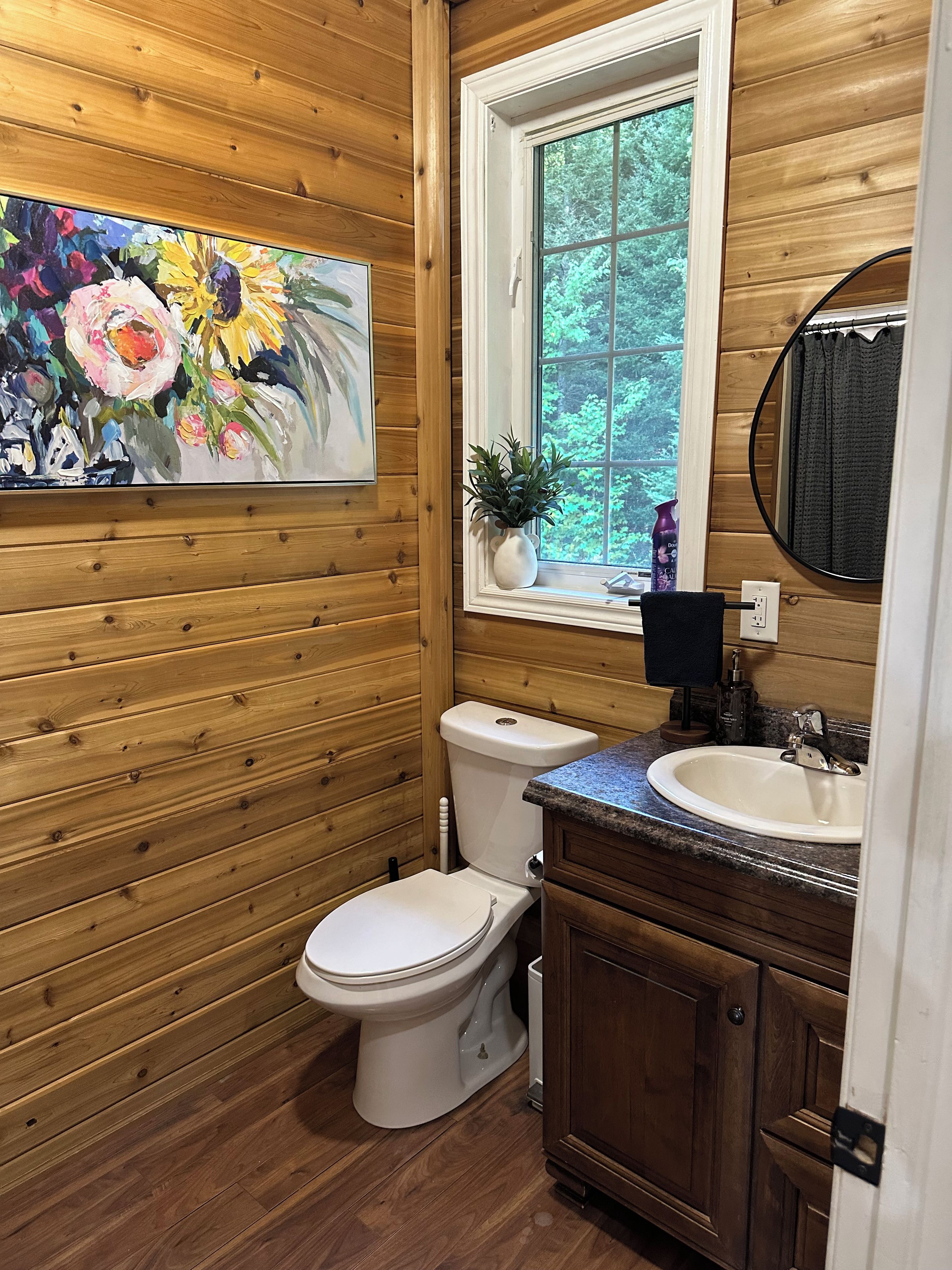 A bathroom with wooden paneling, a white toilet, a sink with a granite countertop, a window with a view of greenery, and a painting of flowers.