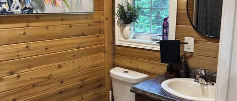 A bathroom with wooden paneling, a white toilet, a sink with a granite countertop, a window with a view of greenery, and a painting of flowers.