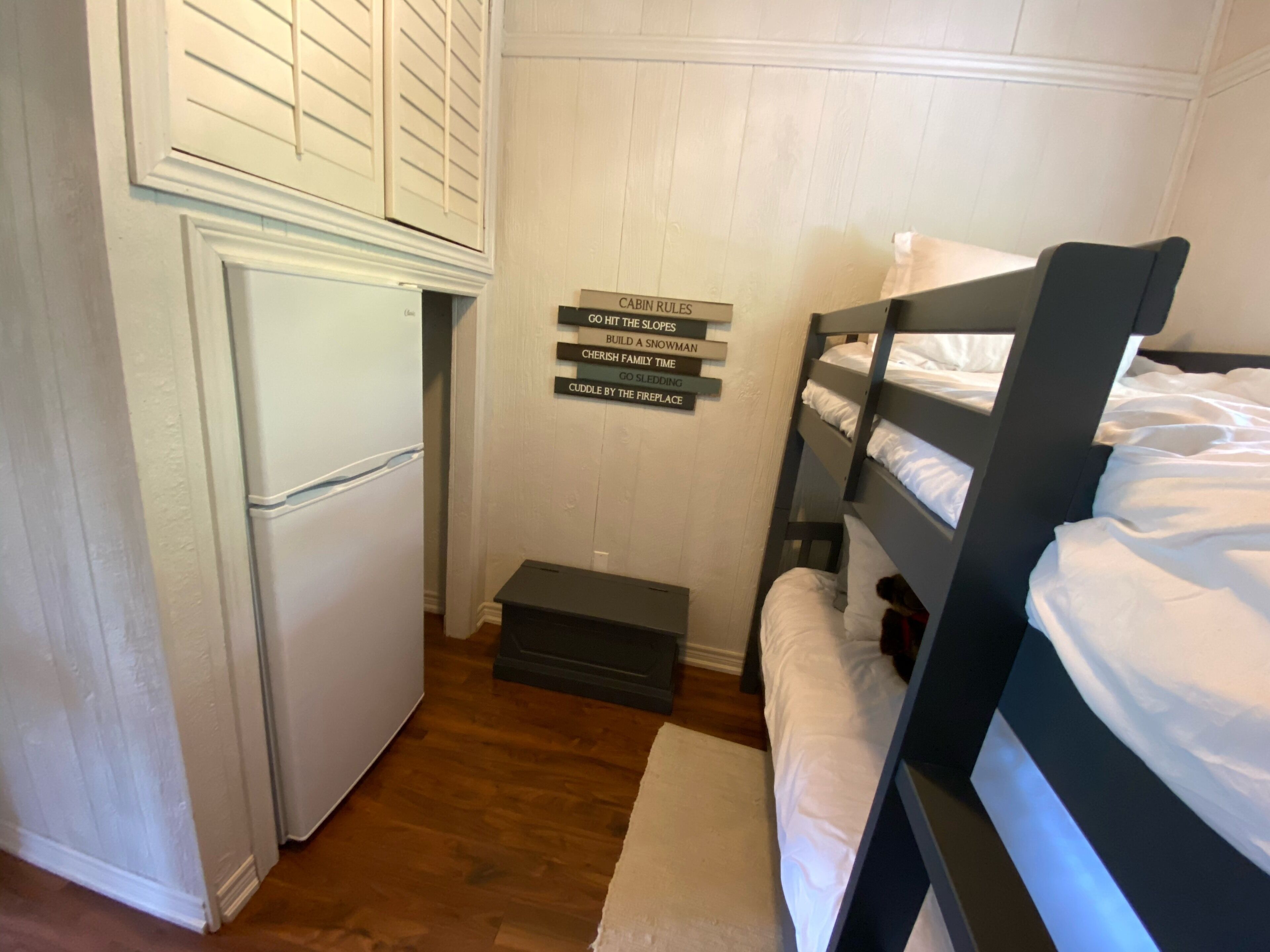 Quarto compacto com beliche, um pequeno frigorífico e um letreiro na parede com várias frases.