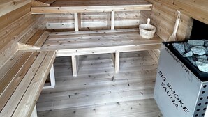 Une salle de sauna avec des bancs en bois et un banc au sol. On y trouve un seau en bois et une cuillère en bois sur une étagère. Une pancarte indique : « ATTENTION, POÊLE CHAUD, FORTE PRODUCTION DE VAPEUR, SAUNA ».