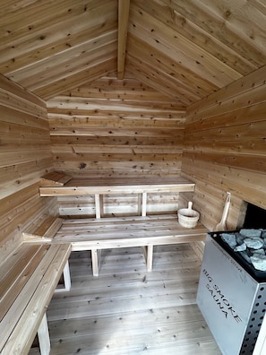 Un cuarto de sauna con bancos de madera y un banco en el suelo. Hay un balde de madera y una cuchara de madera en un estante. Un letrero dice 'CAUTION HOT STOVE BIG SMOKE SAUNA'.