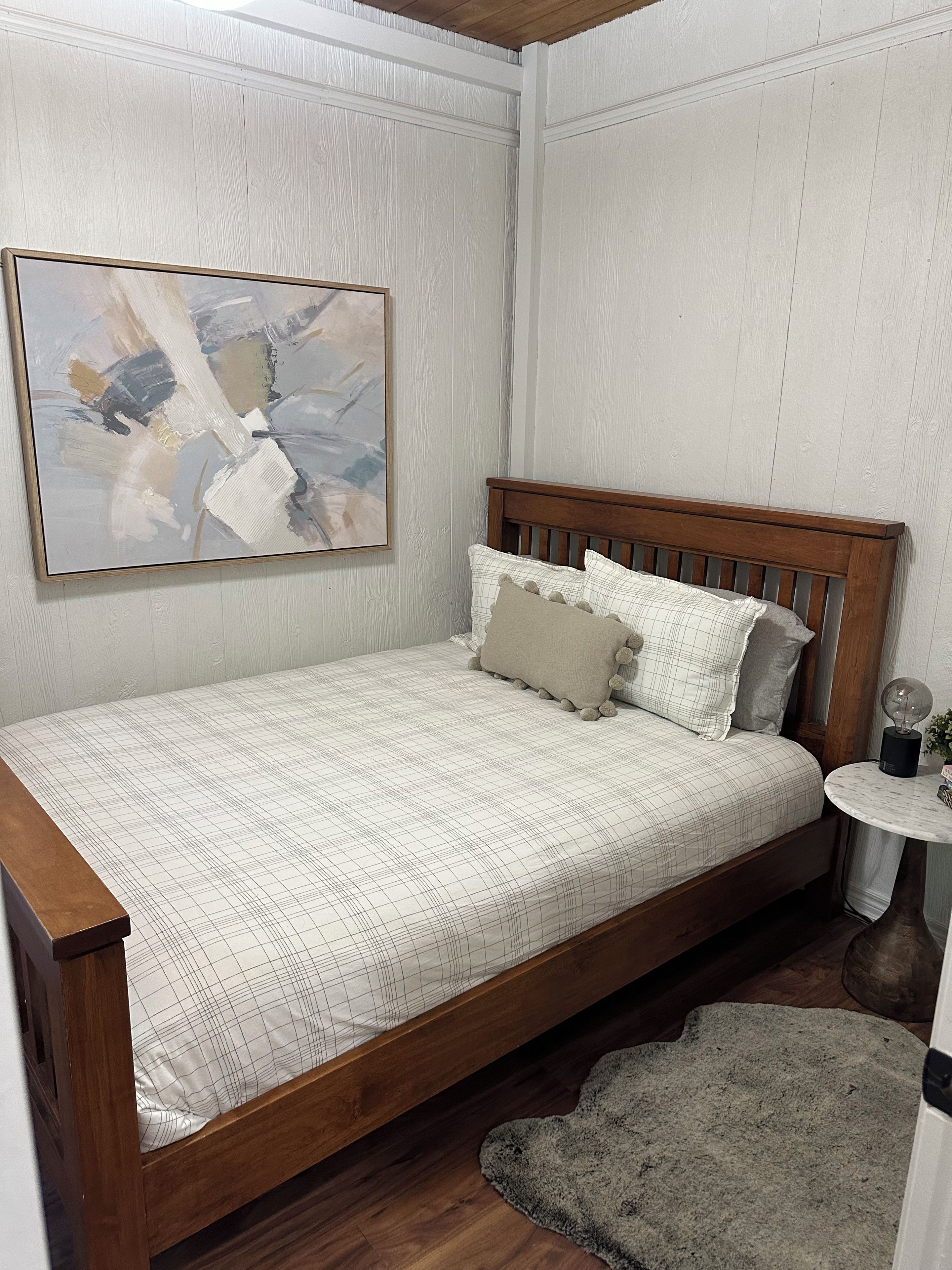 Ein Schlafzimmer mit einem hölzernen Bett, einer weißen Wand mit einem Gemälde und einem kleinen runden Tisch mit einer Lampe.