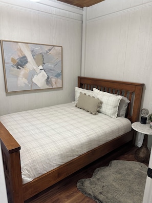 A bedroom with a wooden bed, a white wall with a painting, and a small round table with a lamp.