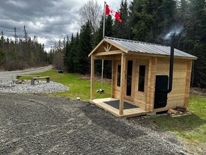 A small wooden cabin with a metal roof, a flagpole with a Canadian flag, and a gravel area with a bench and picnic table. - Cozy 3BR cottage on 100acres with hiking trails, firepit, woodstove & sauna (Burks Falls)