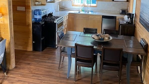 Een houten chalet van binnen, met een eettafel, stoelen, een keuken met kastjes en een magnetron, en een waterdispenser.