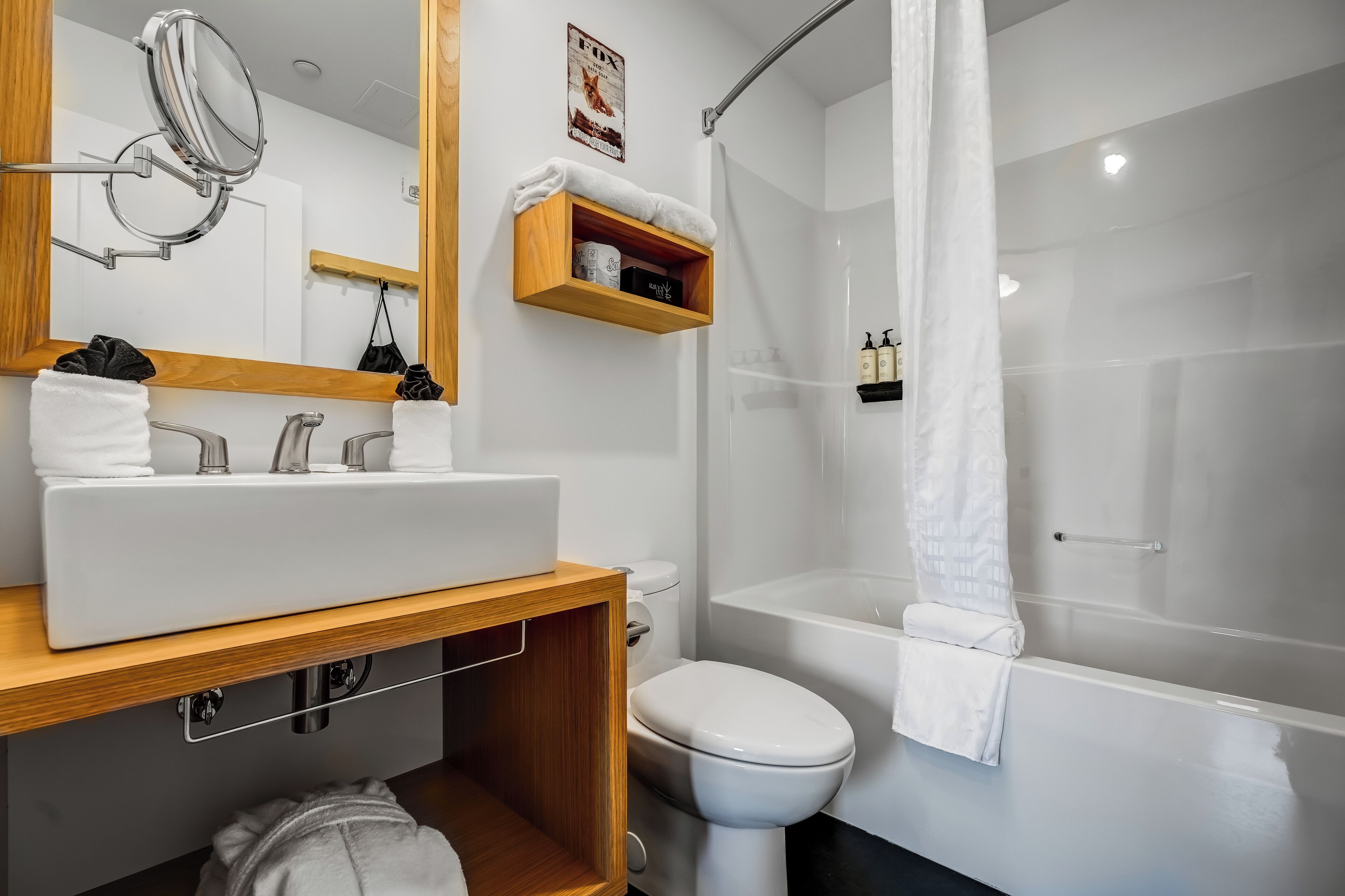 A modern bathroom with a white sink, toilet, and bathtub. There is a wooden shelf with toiletries and a towel hanging on the wall.