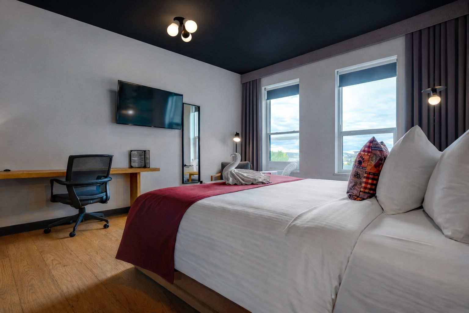 Una habitación de hotel con cama, escritorio, silla, televisor y ventanas amplias.