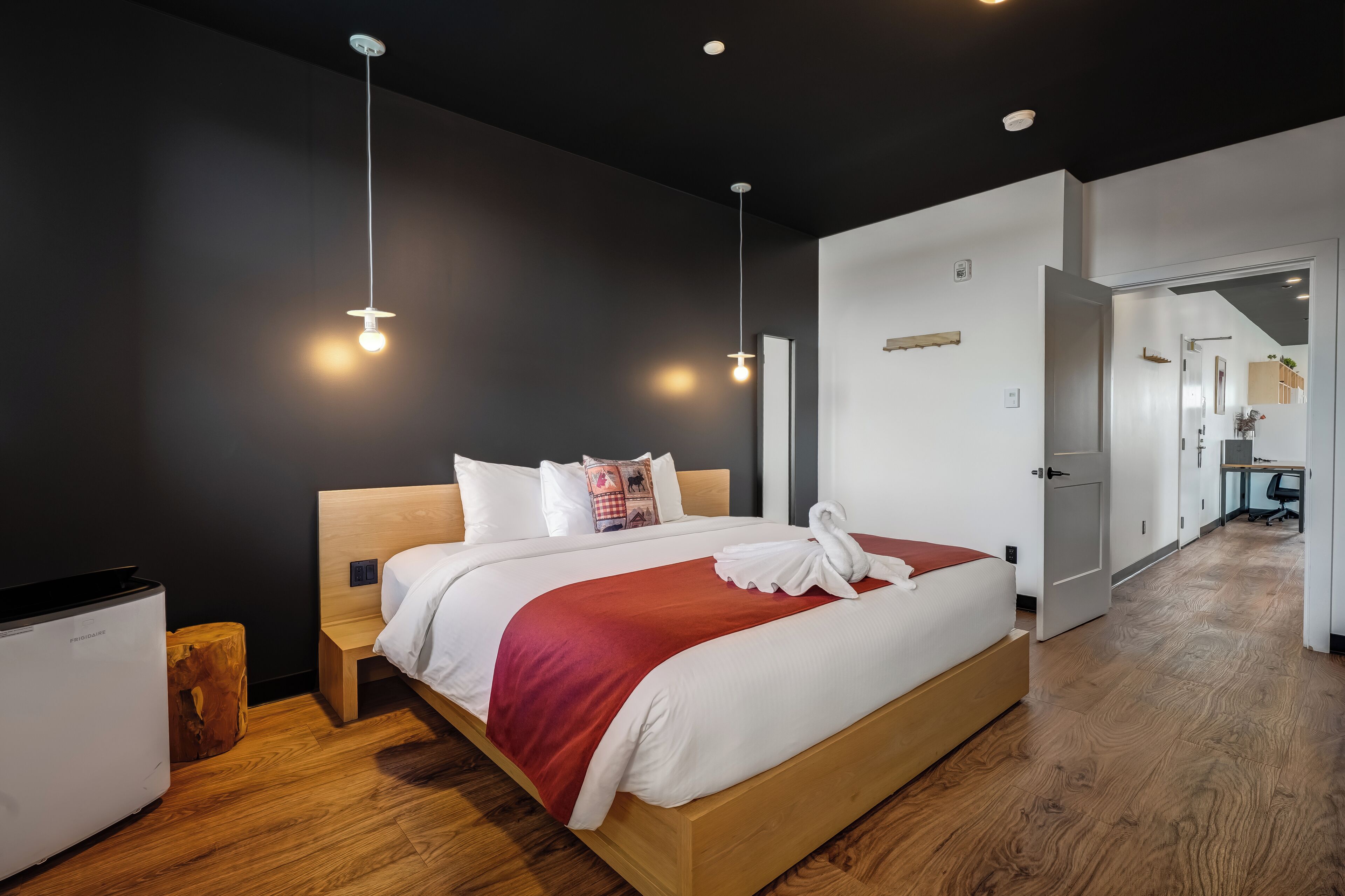 Una habitación de hotel moderna con una cama grande, cabecera de madera y una almohada decorativa roja. Se observa un mesita de noche de madera con una lámpara, un taburete pequeño de madera y un minibar blanco. La habitación tiene paredes oscuras y piso de madera.