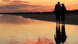 A couple standing on a beach at sunset with a reflection on the wet sand.