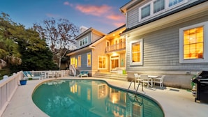 Una piscina con un solárium, un porche cubierto con mobiliario exterior y una casa de dos pisos con balcón.