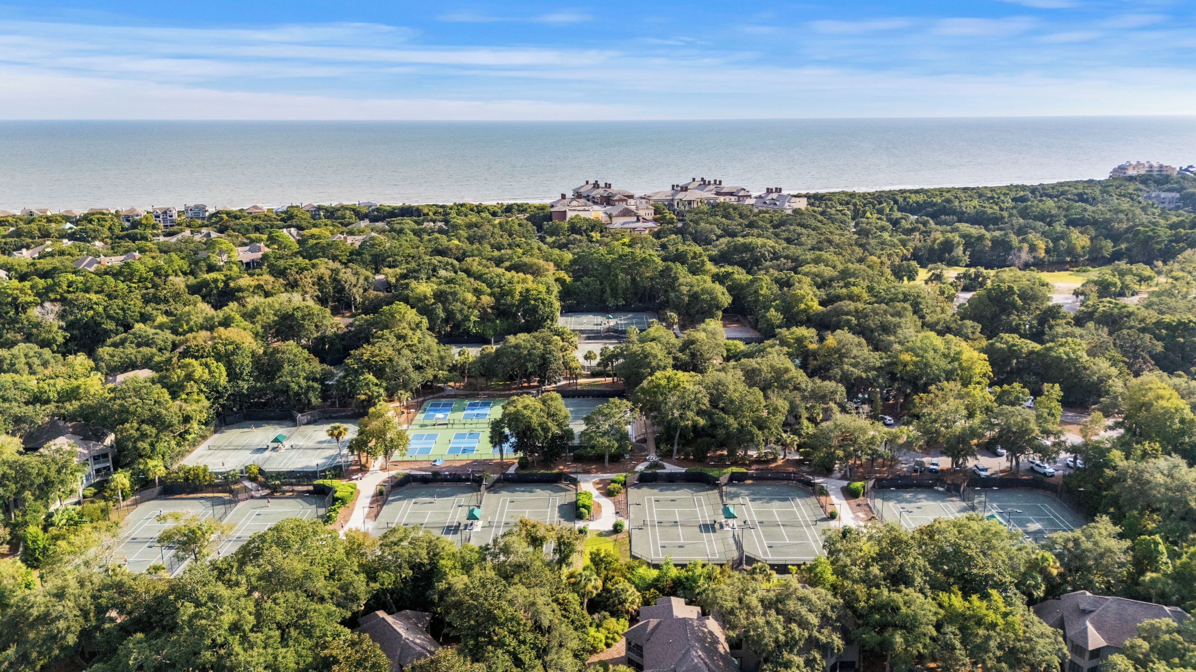 An aerial view of a residential area with multiple tennis courts surrounded by dense greenery, a clear view of the sea, and a distant cluster of buildings.