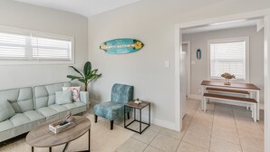 Un salon avec un canapé vert clair, un fauteuil bleu, une table basse et une petite table d’appoint. Un panneau décoratif est accroché au mur, et une porte donne accès à une autre pièce.