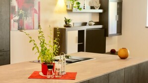 Eine moderne Küche mit einer zentralen Kochinsel, einem Spülbecken und einem Herd. Auf der Insel stehen Topfpflanzen, eine Vase und ein kleiner Tisch mit einer Kerze.
