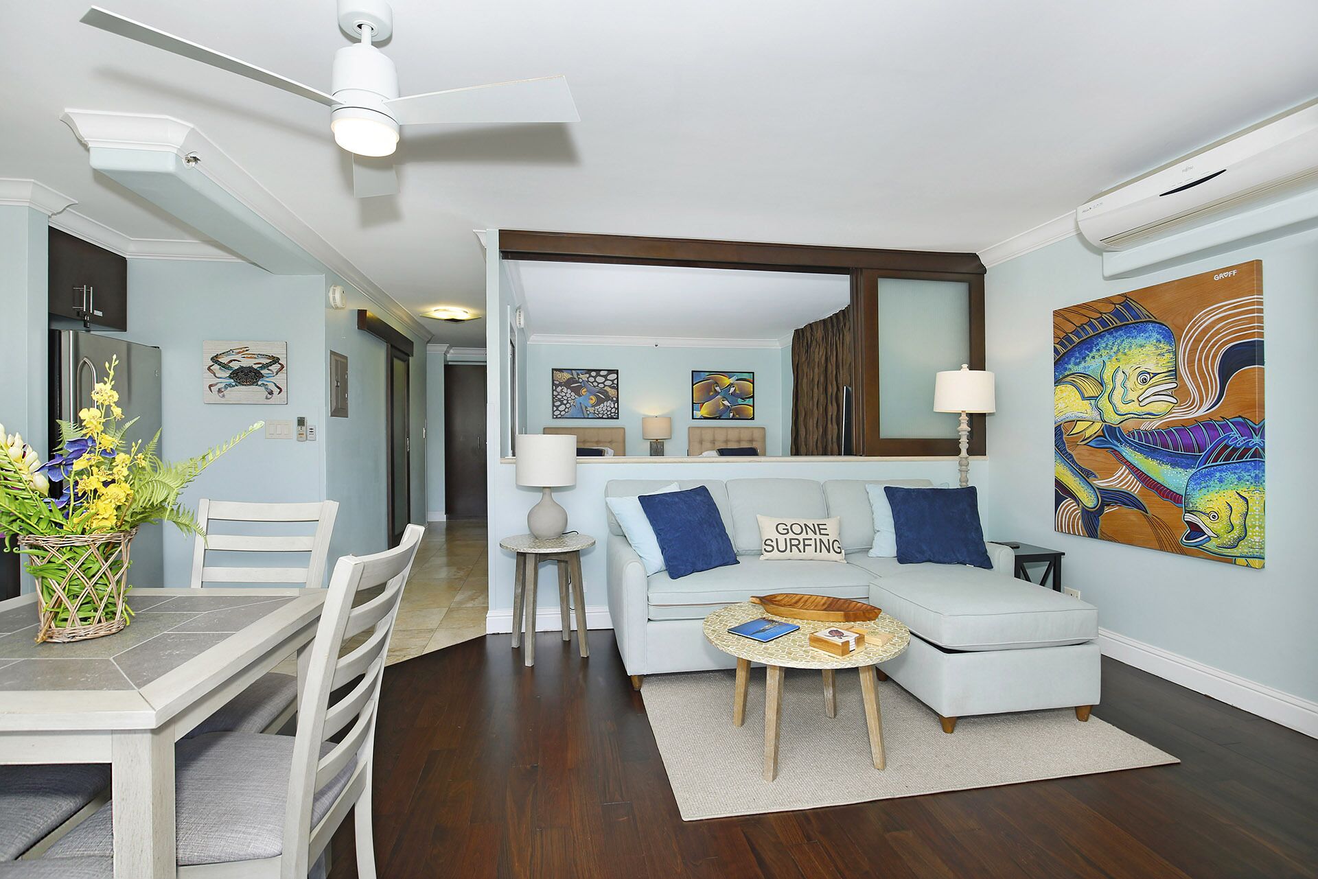 A modern living room with a light blue sofa, a dining table with chairs, a ceiling fan, and artwork on the walls.