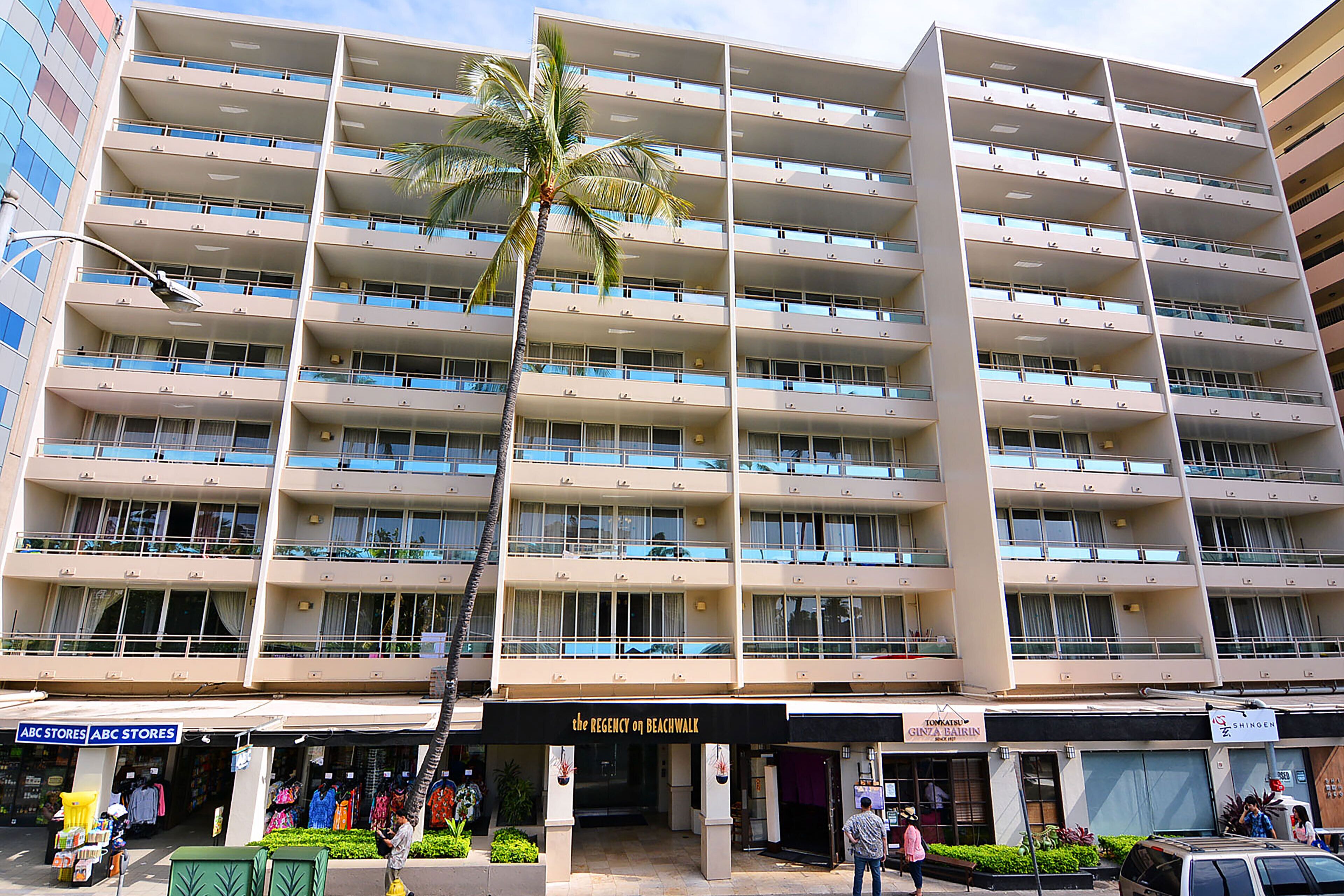 Un hotel de varios pisos con balcones, una palmera y tiendas, incluyendo 'ABC Stores' y 'The Regency on Beachwalk'.