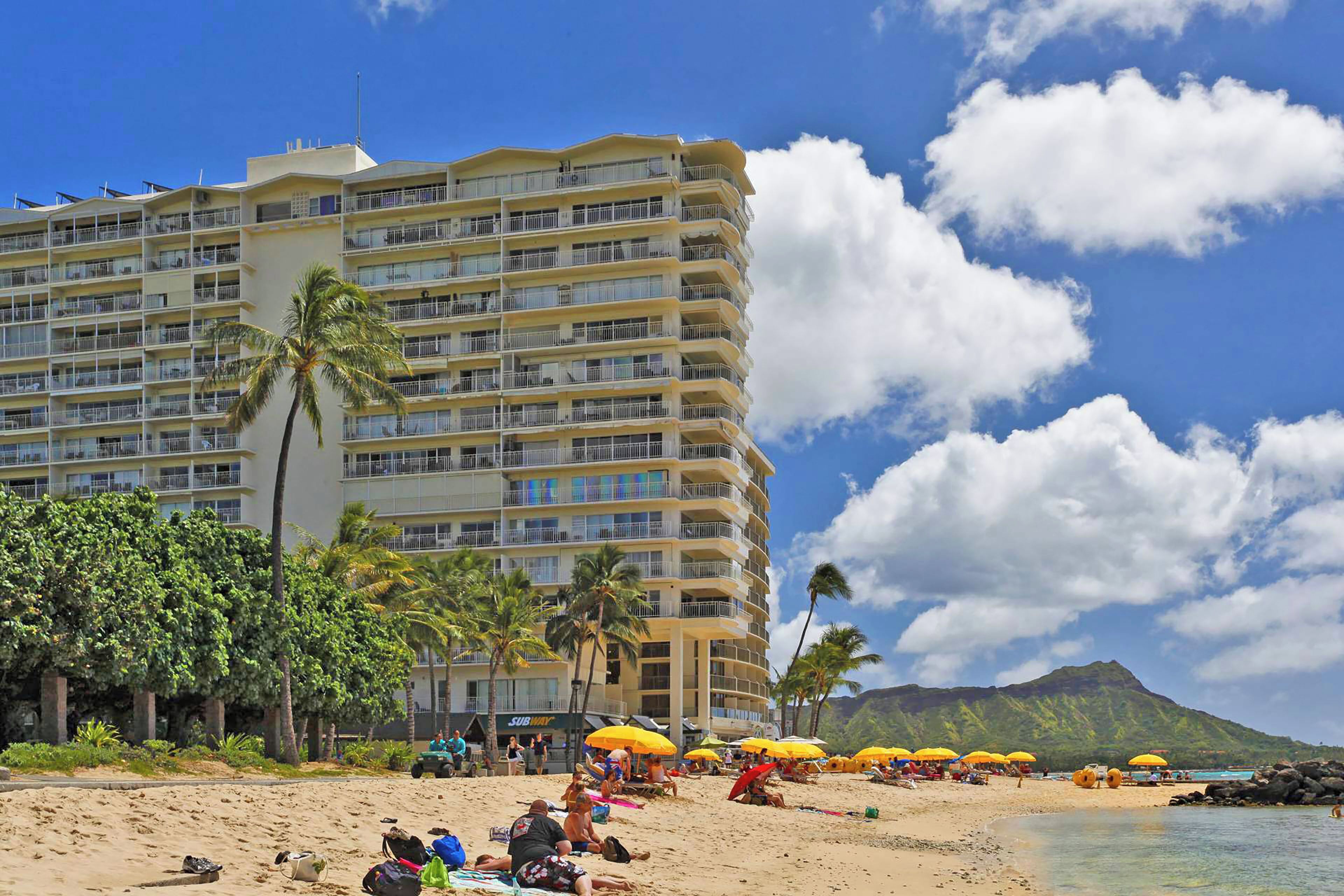 Un hotel frente a la playa con varios balcones, palmeras y sombrillas.