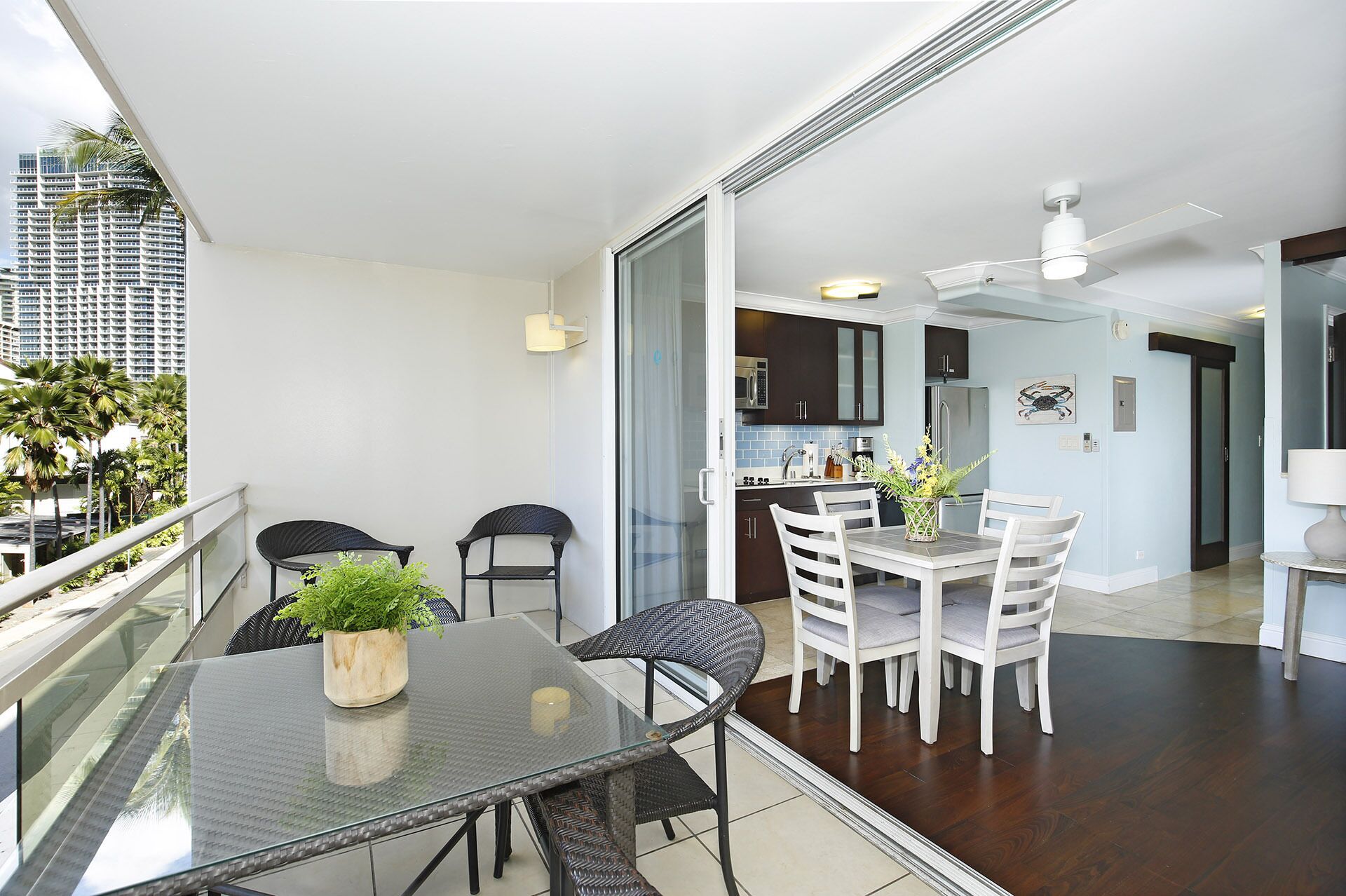 A modern apartment with a balcony, a dining area with a glass table, and a kitchen visible through an open door.