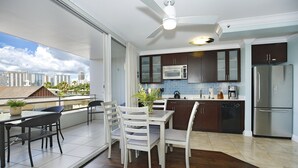 A modern kitchen with dark cabinets, a microwave, and a refrigerator. There is a dining area with a table and chairs, and a balcony with a view of the city.