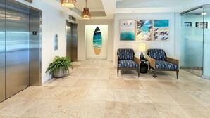 A lobby with a surfboard mural, two patterned chairs, a potted plant, and a modern elevator.
