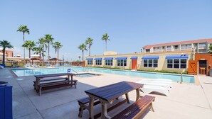 A pool area with wooden benches and lounge chairs, surrounded by palm trees and a building with blue awnings. - Relaxing , Great Location, 3 Minute Walk To The Beach 2 Bedroom Condo by RedAwning (South Padre Island)