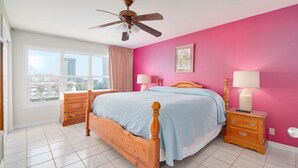 A bedroom with a wooden bed, two bedside tables with lamps, a ceiling fan, a window with curtains, and a pink wall. - Relaxing , Great Location, 3 Minute Walk To The Beach 2 Bedroom Condo by RedAwning (South Padre Island)