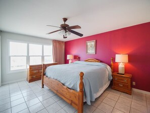 Un dormitorio con cama de madera, dos mesitas de noche, ventilador de techo y ventana con cortinas.