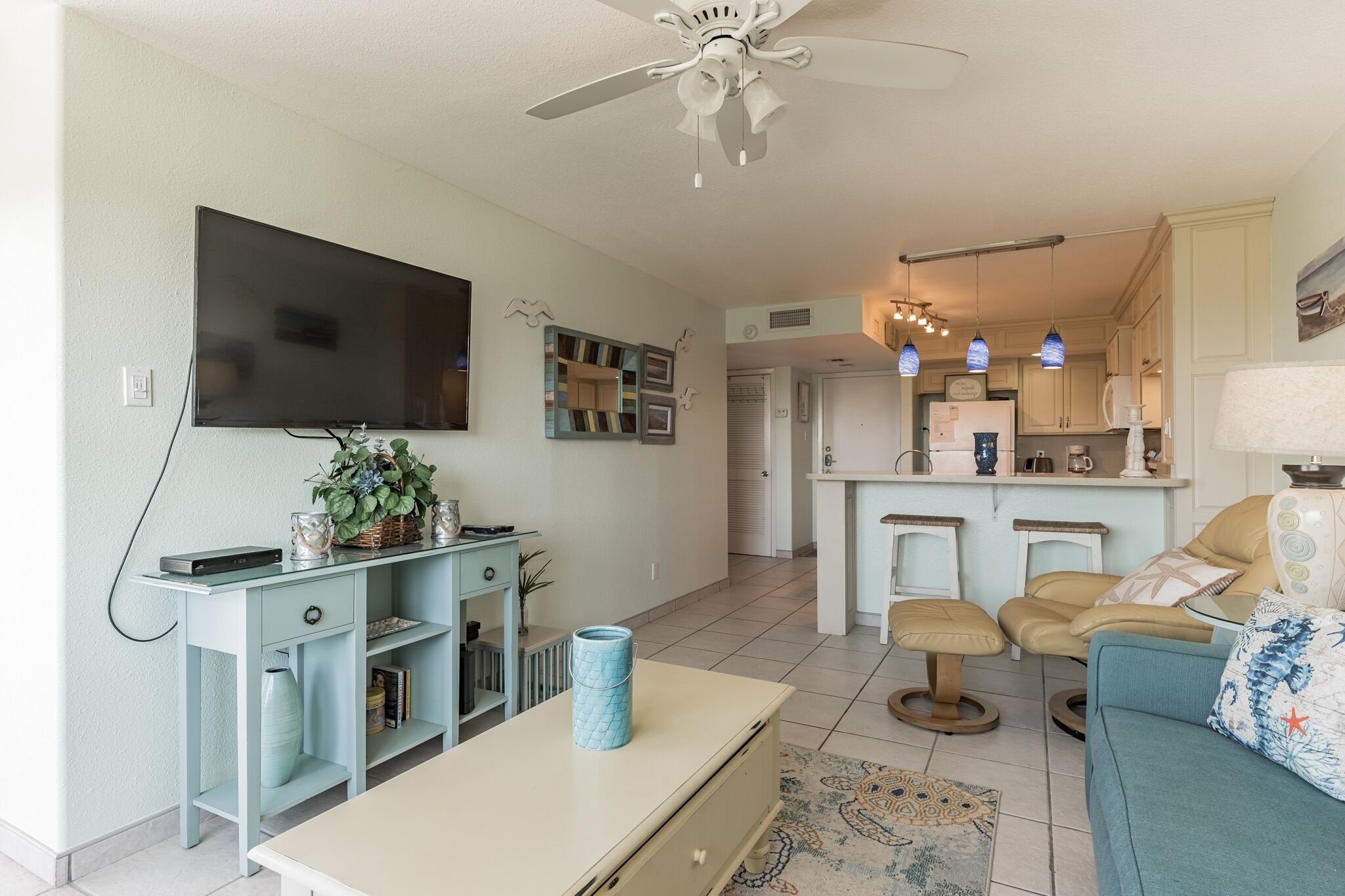 A modern living room with a flat-screen TV, light blue furniture, a ceiling fan, and a kitchen area with pendant lights and a microwave.