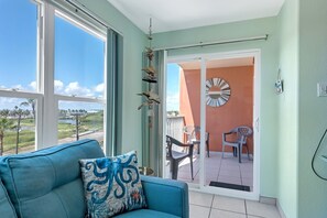 Un balcón con vistas a una playa y palmeras, un sofá azul con un cojín decorativo y un espejo redondo en la pared.