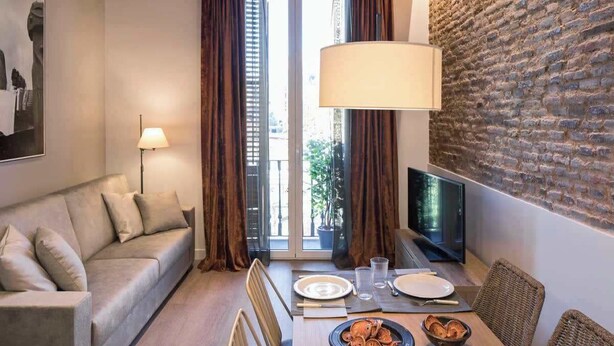 A modern living room with a sofa, dining table, and a TV. The room features exposed brick walls and wooden beams on the ceiling. - Pau Claris | Design 1BR home near Paseo de Gracia (Barcelona)