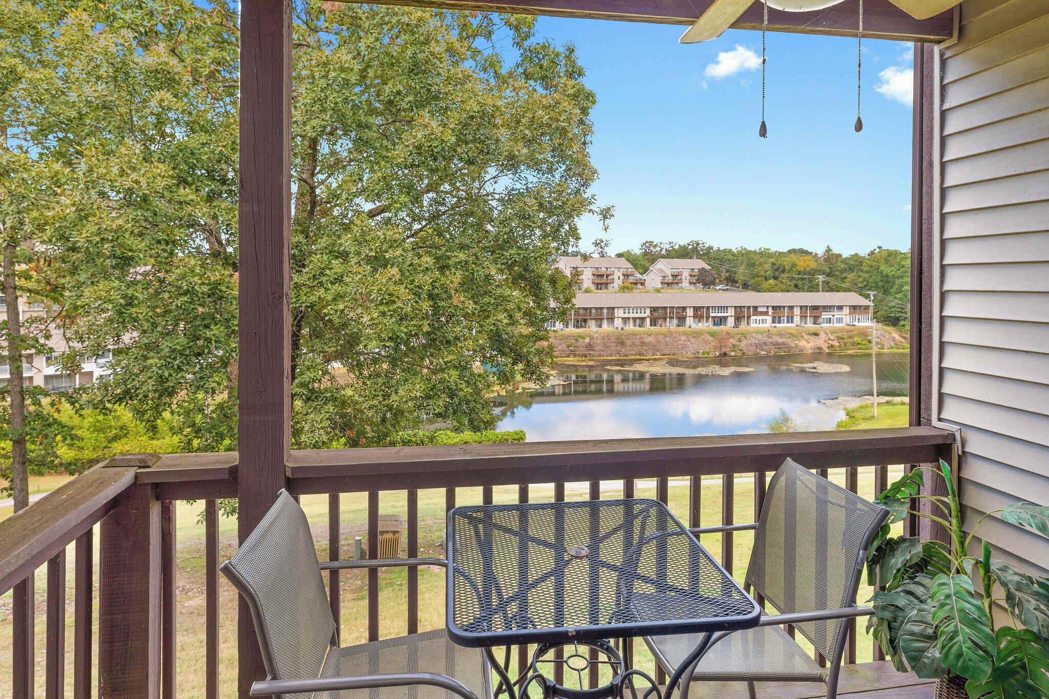 Une terrasse en bois avec deux chaises en métal et une table, d’où l’on a vue sur un lac, avec des bâtiments en arrière-plan.