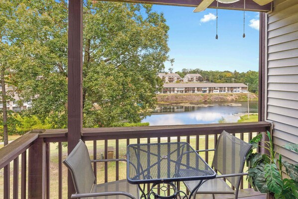 A wooden deck with two metal chairs and a table, overlooking a lake with buildings in the background.