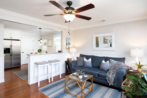 A modern living room with a dark blue sofa, a coffee table, and a kitchen area with white cabinetry and a stainless steel refrigerator.