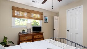 A bedroom with a bed, a dresser, a television, a window with blinds, a door, and a plant. - Palm Key - Cozy Coastal Charm (Charleston)