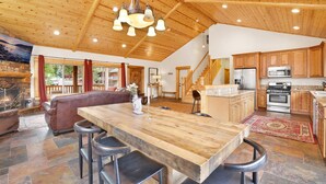 A spacious living area with a wooden ceiling, a stone fireplace, a large wooden dining table with bar stools, and a kitchen with wooden cabinets and stainless steel appliances. - Eagle Ridge Views #1648 (Big Bear Lake)