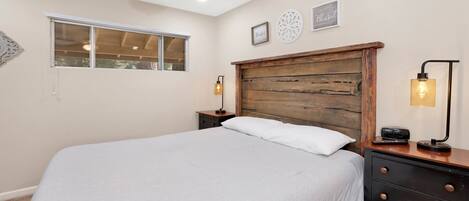 A bedroom with a wooden headboard, a large bed, two bedside tables with lamps, a window, and wall decorations.
