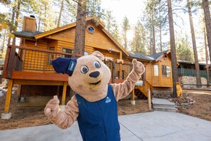 Een beer mascotte staat voor een houten chalet met een terras en een trap naar de ingang.