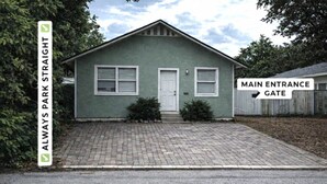 A single-story building with a green facade, a white door, and a brick driveway. A sign indicates the main entrance gate.