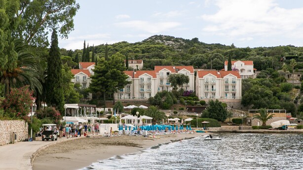 A coastal hotel with multiple balconies overlooking the beach, a sandy shoreline, and a hill with dense greenery in the background. - Apartments Maria Koločep - Two Bedroom Apartment with Terrace and Garden View (Kolocep)