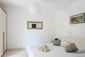 Une chambre à coucher comprenant un lit, des oreillers, une table de chevet, une commode, un luminaire suspendu, un tableau encadré et une armoire.