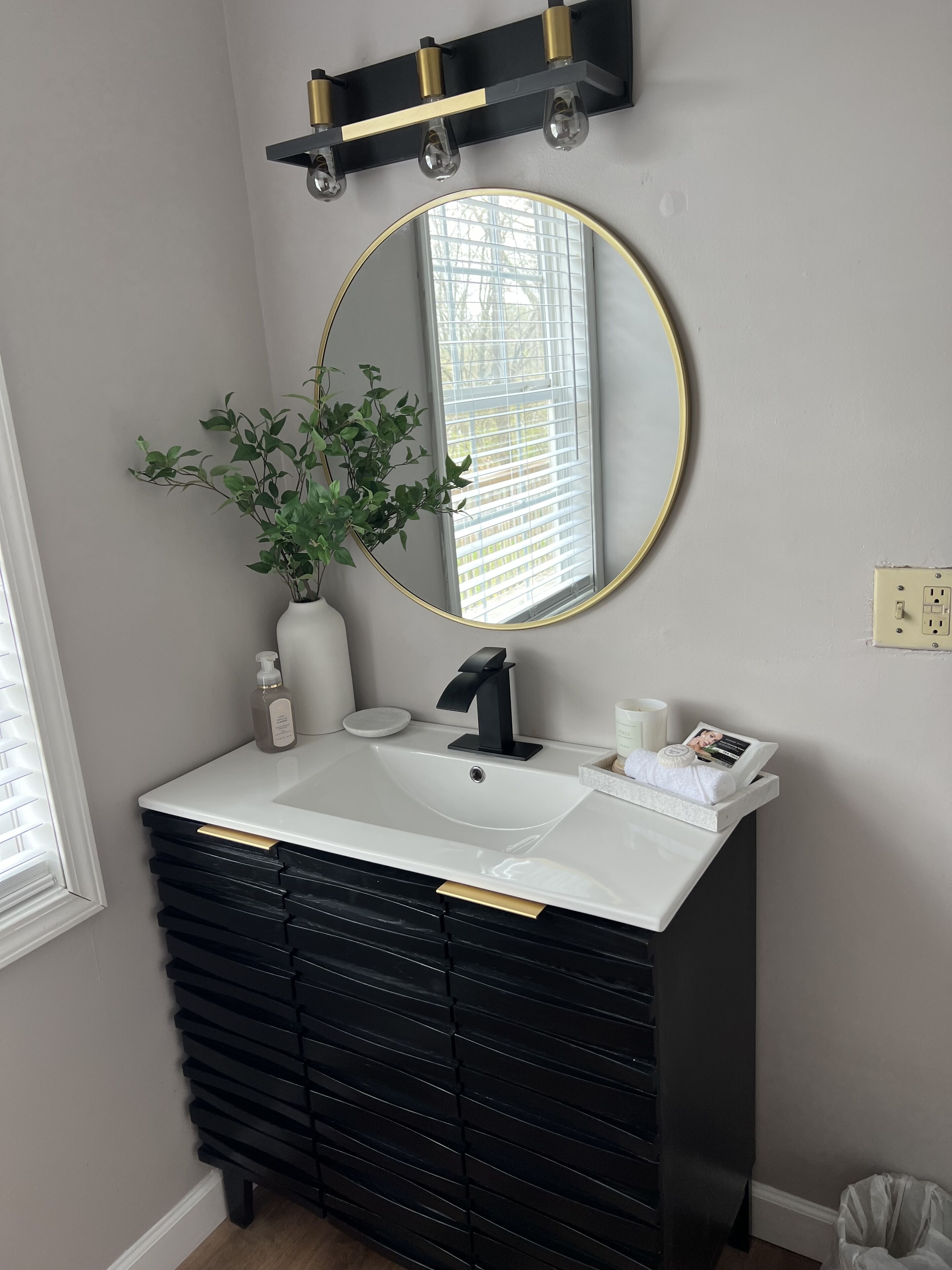 Une salle de bain avec un lavabo blanc, un meuble-lavabo noir, un miroir rond avec un cadre doré, une plante en pot et une fenêtre avec des stores.